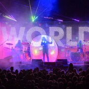 Un chanteur dans une bulle entouré de deux batteurs, deux guitaristes et deux claviéristes sur une scène devant une foule.