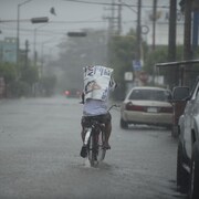 Un homme circule sur un vélo dans une rue recouverte par quelques centimètres d'eau. Le temps est lourd et il pleut.