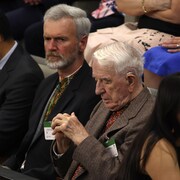 Un vieil homme assis au milieu d'autres personnes, les yeux fermés.
