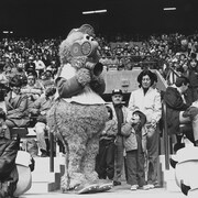 Entourée de la foule au stade olympique de Montréal, la mascotte Youppi! tient une caméra de Radio-Canada.