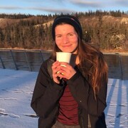 Une jeune femme tient un gobelet en plastique dans les mains. Elle pose devant une rivière. Il y a des arbres de l'autre côté de la rive.