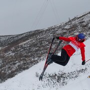 Une athlète en ski effectue un saut sur un parcours de demi-lune. On voit les montagne derrière elle.