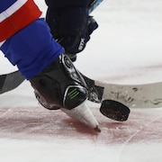 Deux bâtons de hockey se disputent la rondelle entre des patins.