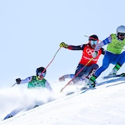 Des skieurs se suivent de près sur un parcours. Le meneur a un dossard jaune et un uniforme bleu.