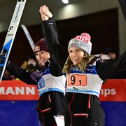 Deux skieuses sourient et lèvent leurs skis en l'air pour saluer la foule.