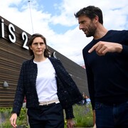 Deux personnes discutent devant un immeuble avec accrochées au mur les lettres Paris 2024.