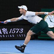 Un joueur de tennis tente de frapper une balle de coup droit, du bout de son bras. On voit qu'il est 3h55 du matin sur l'horloge du terrain. 