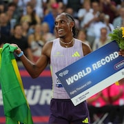 Alison dos Santos tout sourire avec un drapeau du Br&eacute;sil dans une main et une affiche de record du monde dans l'autre.