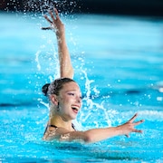 Audrey Lamothe dans la piscine en train de performer, avec le bras gauche levé vers le haut, le bras droit tendu vers l'avant et un grand sourire.