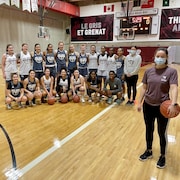 Une femme se tient avec un masque quelques pas devant son équipe de basketball qui prend la pose avant un entraînement.