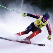 Un skieur casqué, de face, dévale une pente, les bras écarté, avec une combinaison de l'équipe canadienne.
