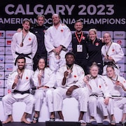 Des judokas souriants et leur équipe de soutien montrent leurs médailles sur un podium devant un mur de commanditaires. 