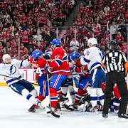 Des joueurs de hockey en blanc et en rouge se chamaillent près d'un but sous le regard des arbitres.