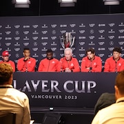 Huit hommes, vêtus du même survêtement rouge, sont assis à une table et répondent aux questions des journalistes en conférence de presse. 