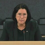 Une femme habillée en civil, avec des lunettes, répond à des questions derrière un micro. On voit des bancs vides derrière elle. 