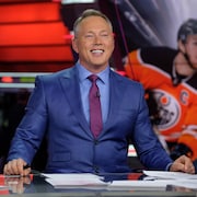 Un homme en complet et cravate est assis derrière un bureau. Il sourit et une image d'un hockeyeur se trouve derrière lui.