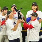 Un homme coiffé d'une casquette tient un trophée alors que des joueurs de baseball l'applaudissent.