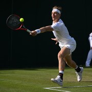 Un joueur de tennis, tout de blanc vêtu, s'étire pour frapper une balle du revers avec sa raquette rouge sur un terrain de gazon. 