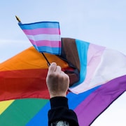 Une main tient un petit drapeau aux couleurs associées aux personnes transgenres et derrière flotte un gros drapeau arc-en-ciel.