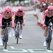 Trois cyclistes dans le maillot rose de l'équipe EF sur leurs vélos de contre-la-montre, le visage crispé par l'effort, approchent de la ligne d'arrivée. 