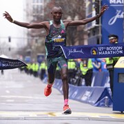 Un homme lève les bras et casse le ruban à l'arrivée d'un marathon. Un chronomètre indique son temps: 2 min 05 s 53