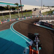 Une monoplace F1 négocie une chicane sur un circuit entouré de murets de protection, on voit des palmiers et une autoroute en arrière-plan avec des voitures qui roulent. 