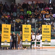 Actuels et anciens joueurs des Broncos de Humboldt, en arrière-plan, regardent les bannières rendant hommage aux victimes de la tragédie.