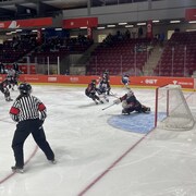 Une gardienne de but devant son filet qui réalise un arrêt devant un joueur adverse sous les regards d'un défenseur et de l'arbitre sur la patinoire.
