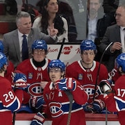 Des joueurs de hockey, l'air sérieux au banc de leur équipe, écoutent leur entraineur donner des instructions.