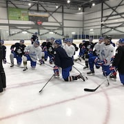 Un entraîneur montre des exercices sur un tableau à des joueurs agenouillés sur la patinoire.