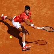 Un joueur de tennis grimace et fait le grand écart pour aller chercher une balle du revers sur un court en terre battue.