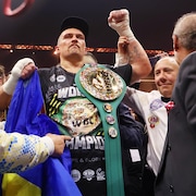 Un boxeur, avec la ceinture de la WBC sur l'épaule, lève le poing gauche et tient le drapeau ukrainien dans la main droite. 