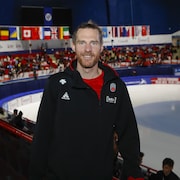 Un homme portant une barbe rousse, vêtu de noir et rouge, prend la pose devant une patinoire. 