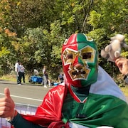 Un partisan du Mexique v&ecirc;tu aux couleurs du pays porte un masque de luchador et l&egrave;ve son pouce devant la cam&eacute;ra. Dans le coin inf&eacute;rieur droit, une photo d'Ilrick Duhamel de Radio-Canada, souriant.