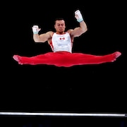 Un gymnaste, vêtu d'un pantalon rouge et d'un chandail blanc, est photographié en faisant le grand écart dans les airs par-dessus la barre fixe. 