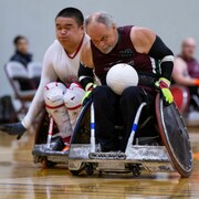 deux joueurs de rugby assis dans un fauteuil roulant, le premier en possession d'un ballon sur une surface de jeu.