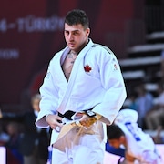Un judoka en judogi orné d'une feuille d'érable a les mains sur sa ceinture noire et regarde devant lui.