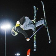 Rachael Karker effectue une figure où elle a la tête vers le sol, tout en attrapant son ski droit de la main droite.