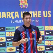 Il lève les pouces en l'air et sourit pour les caméras devant un grand panneau avec le logo du FC Barcelone et ceux de commanditaires.