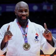 Un judoka souriant lève ses deux index en signe de victoire, avec une médaille d'or au cou.