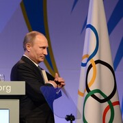 Un homme range un objet dans la poche intérieure de son veston en marchant devant le drapeau olympique.