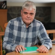 Un homme, assis, regarde la caméra en tenant un dossier dans ses mains.