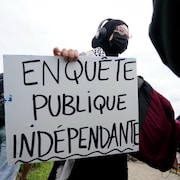 Une manifestante portant un masque tient une pancarte où l'on peut lire «Enquête publique indépendante».