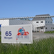 Un bâtiment industriel photographié de l'autre côté d'une clôture de broche qui entoure le terrain. Une affiche identifiant l'usine est accrochée à la clôture.
