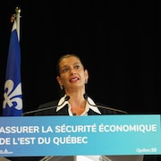Geneviève Guilbault, une femme aux cheveux foncés attachés, vêtue d'une veste de costume noire et d'une chemise blanche, parle avec les bras écartés derrière un pupitre bleu affichant le texte "ASSURER LA SÉCURITÉ ÉCONOMIQUE DE L'EST DU QUÉBEC". 