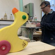Un canard jaune en bois au premier plan. Derrière, un homme est en train d'utiliser une machine à travailler le bois.