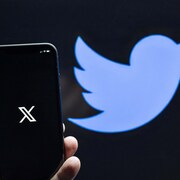 Un main tient un téléphone avec le logo de X devant un mur avec l'ancien logo de Twitter.