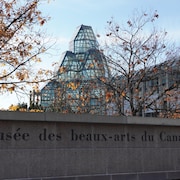 Le nom du musée est écrit sur un muret de briques. En arrière-plan, l'édifice du musée et des arbres avec quelques feuilles oranges.