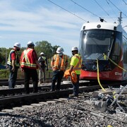 Un groupe de personnes portant des vestes de sécurité et des masques, sur les rails, devant un train léger à l'arrêt.