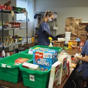 des aliments sur une table et des étagères. Deux jeunes femmes préparent des cartons de nourriture pour les étudiants